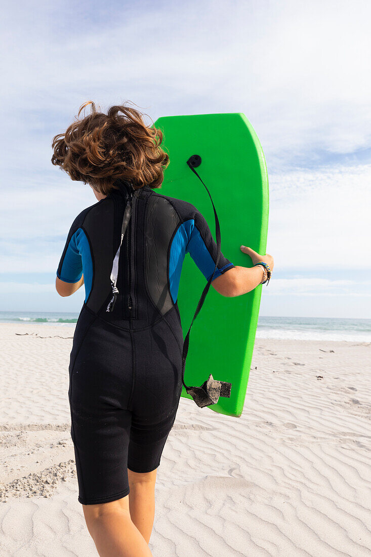 Südafrika, Hermanus, Rückansicht eines Jungen, der mit einem Bodyboard am Strand läuft