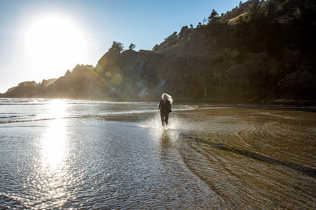 USA, Oregon, Newport, Frau läuft am Sandstrand und spritzt Wasser