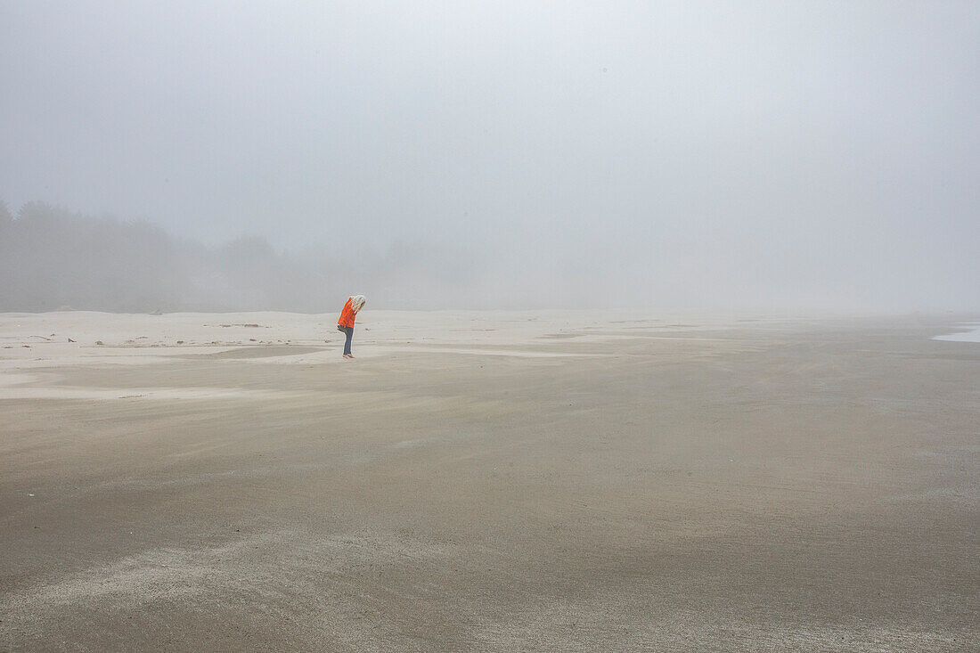 Frau in orangefarbener Jacke steht am nebligen Strand