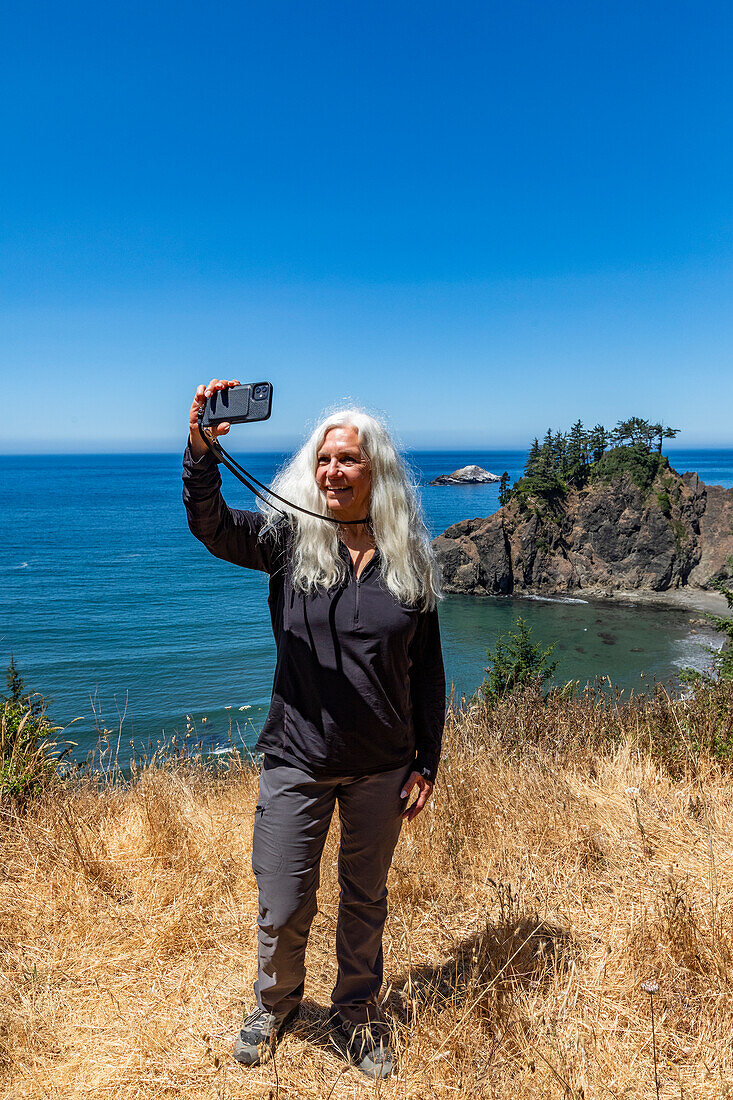USA, Oregon, Brookings, Senior woman taking selfie with coastal landscape in background