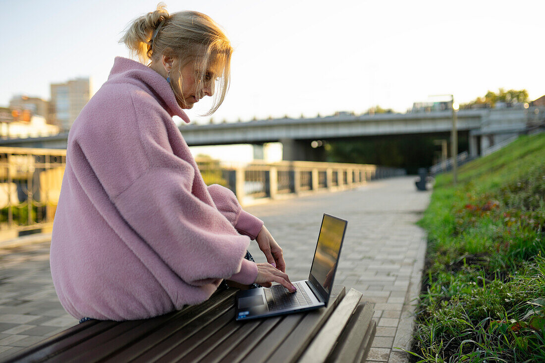 Frau arbeitet an einem Laptop, während sie bei Sonnenaufgang auf einer Bank sitzt