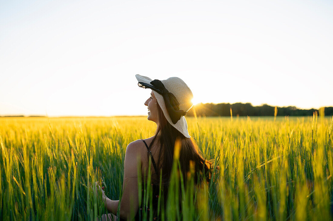 Rückansicht einer Frau mit Strohhut, die bei Sonnenuntergang in einem Feld steht