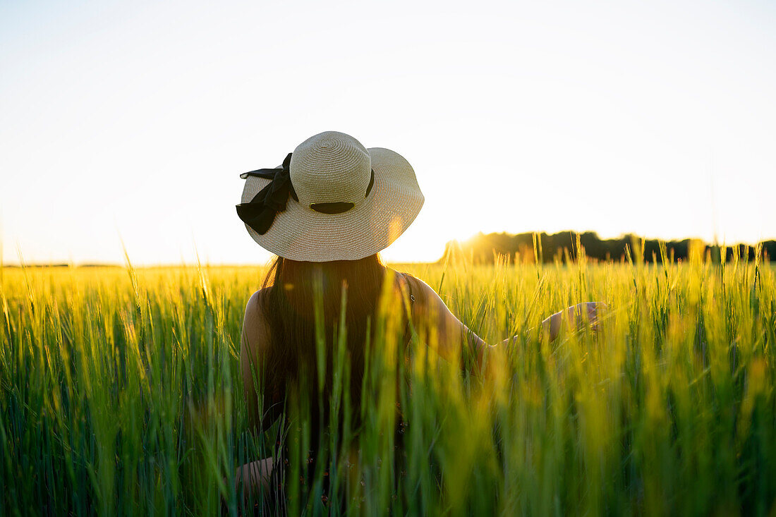 Rückansicht einer Frau mit Strohhut, die bei Sonnenuntergang in einem Feld steht