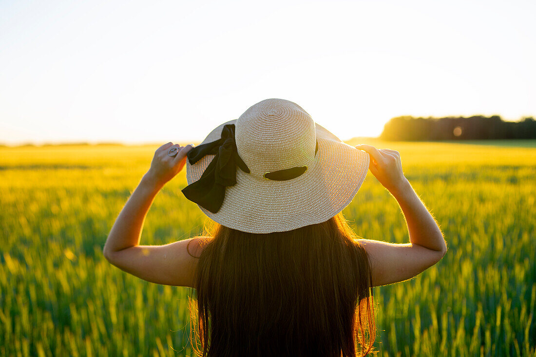 Rückansicht einer Frau mit Strohhut, die bei Sonnenuntergang in einem Feld steht