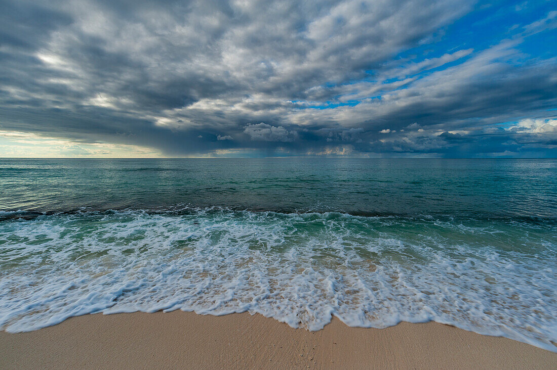 Die Brandung des Indischen Ozeans brandet unter einem dramatischen Wolkenhimmel auf einen Sandstrand. Denis-Insel, Seychellen.