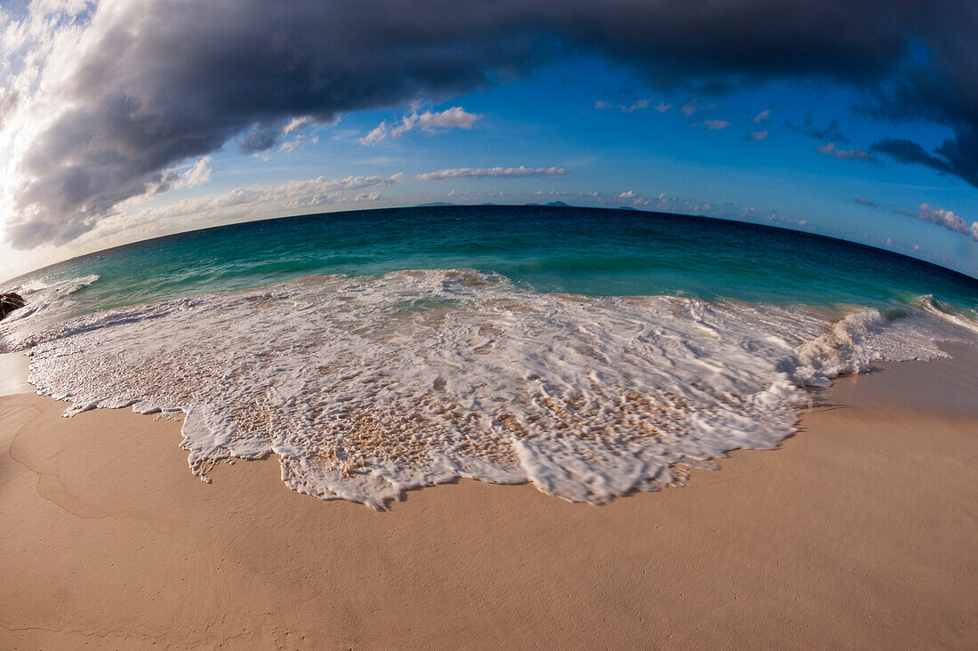 A fisheye lens view of Indian Ocean surf surging onto a sandy tropical beach. Anse Macquereau Beach, Fregate Island, Seychelles.