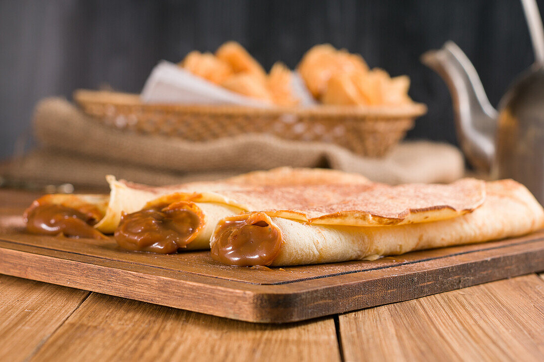 Frisch gerollte Crêpes mit süßer Dulce de Leche-Füllung, serviert auf einem hölzernen Schneidebrett auf einem Tisch mit Wasserkocher in der Küche