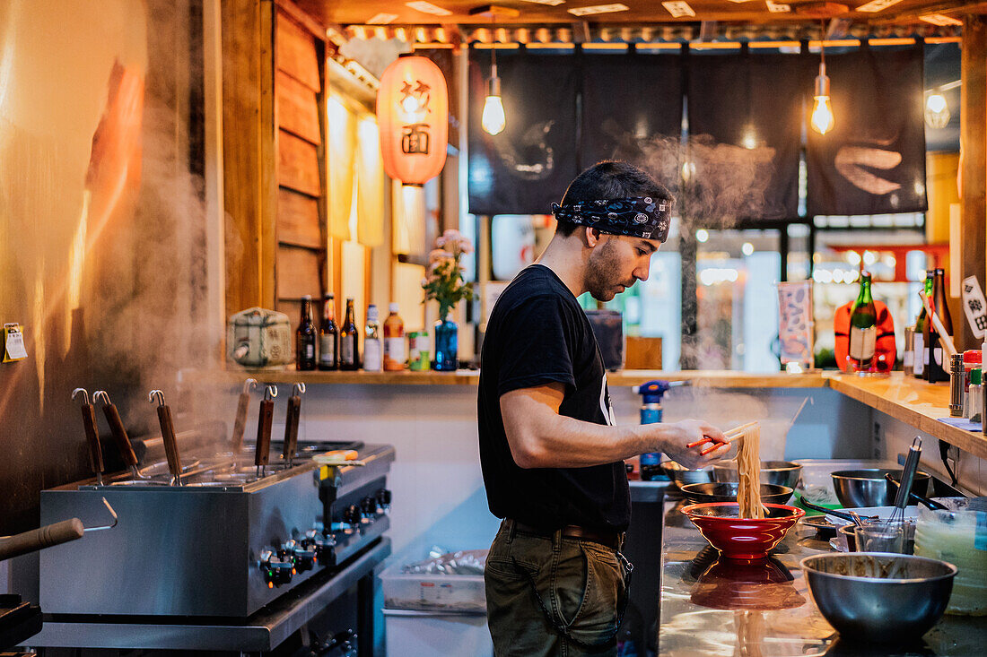 Seitenansicht eines Mannes mit Kopftuch, der an der Theke steht und Ramen in einem modernen asiatischen Café kocht