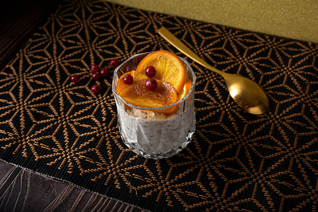 Blick von oben auf ein Glas mit einem Dessert aus Stracciatella-Mousse und Schokoladenspänen, gekrönt von karamellisierten Orangenscheiben und Beeren neben einem Löffel