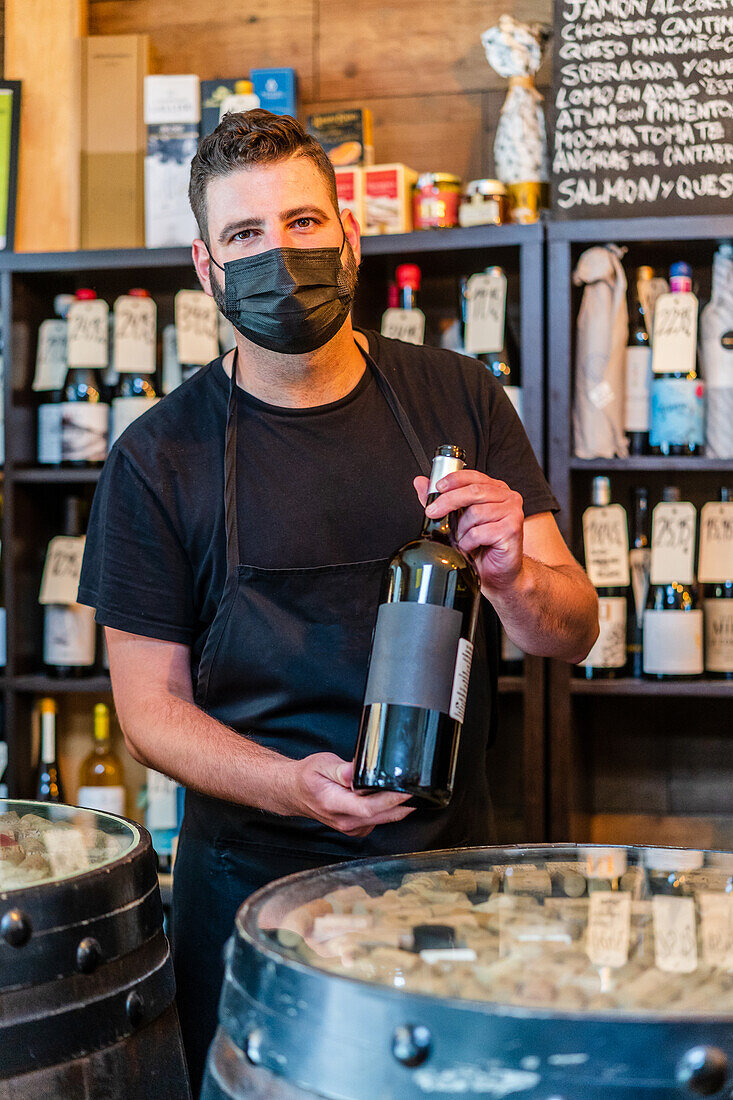 Seriöser bärtiger Barkeeper mit dunklem Haar in schwarzer Schürze steht am Tresen und zeigt eine Flasche Rotwein, während er im Restaurant arbeitet