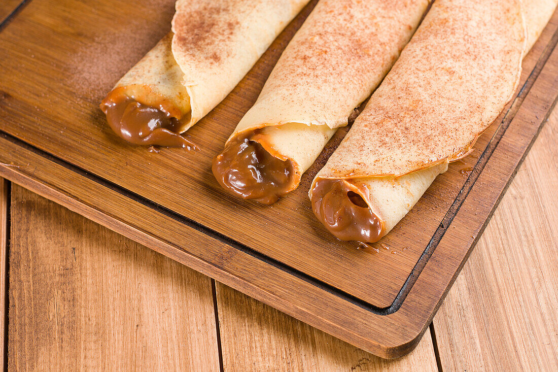 Frische, schmackhafte gerollte Crêpes mit süßer Dulce de Leche-Füllung, serviert auf einem hölzernen Schneidebrett in der Küche (von oben)