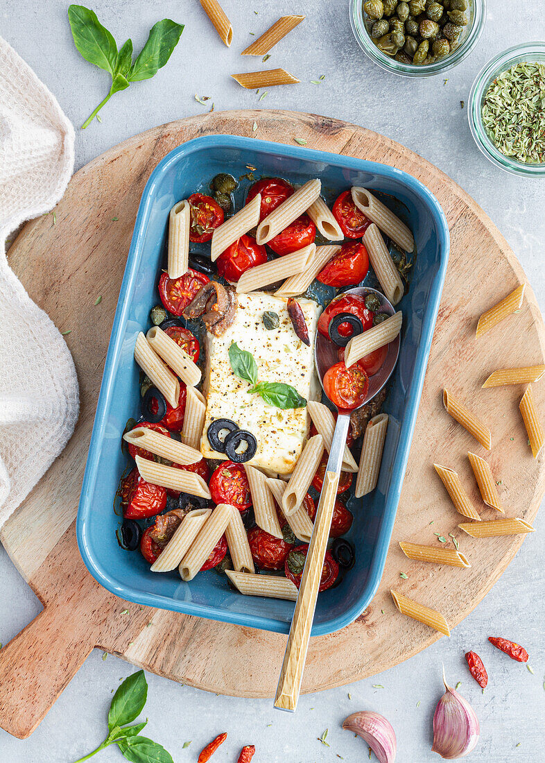 Reife Kirschtomaten mit schwarzen Olivenscheiben und Fetakäse in einer Auflaufform neben ungekochten Penne von oben