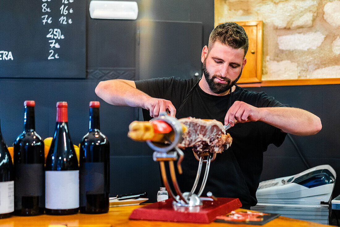 Bärtiger professioneller ethnischer Schneider bereitet leckeren Jamon in einem Restaurant mit Weinflaschen zu