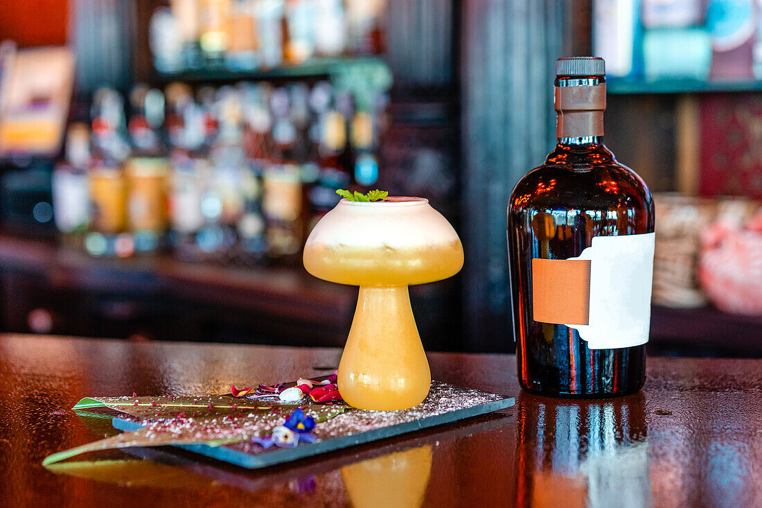 Saurer Cocktail mit Minzblatt in einem pilzförmigen Glas, serviert auf einem Holztresen mit einer Flasche Alkohol in einem Pub