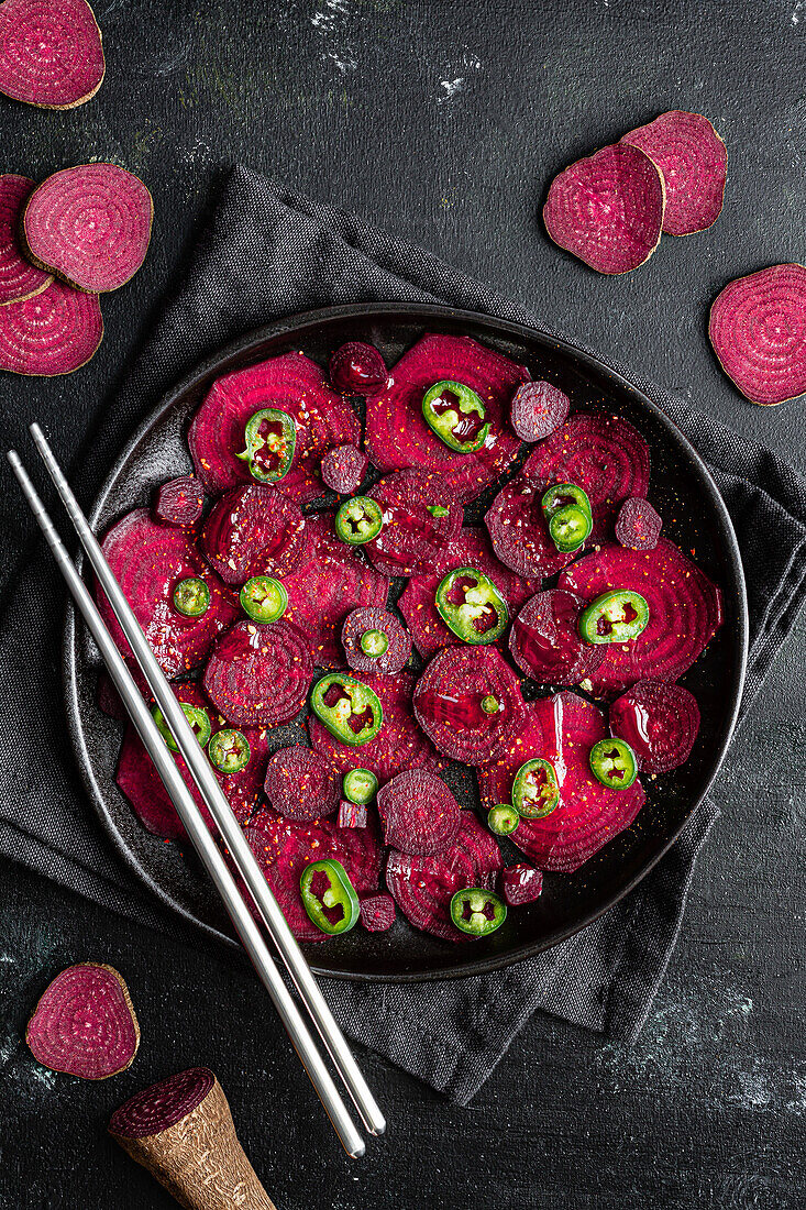 Top view composition of tasty beetroot slices arranged on baking pan with green jalapeno peppers and placed on black towel on kitchen table close to chopsticks