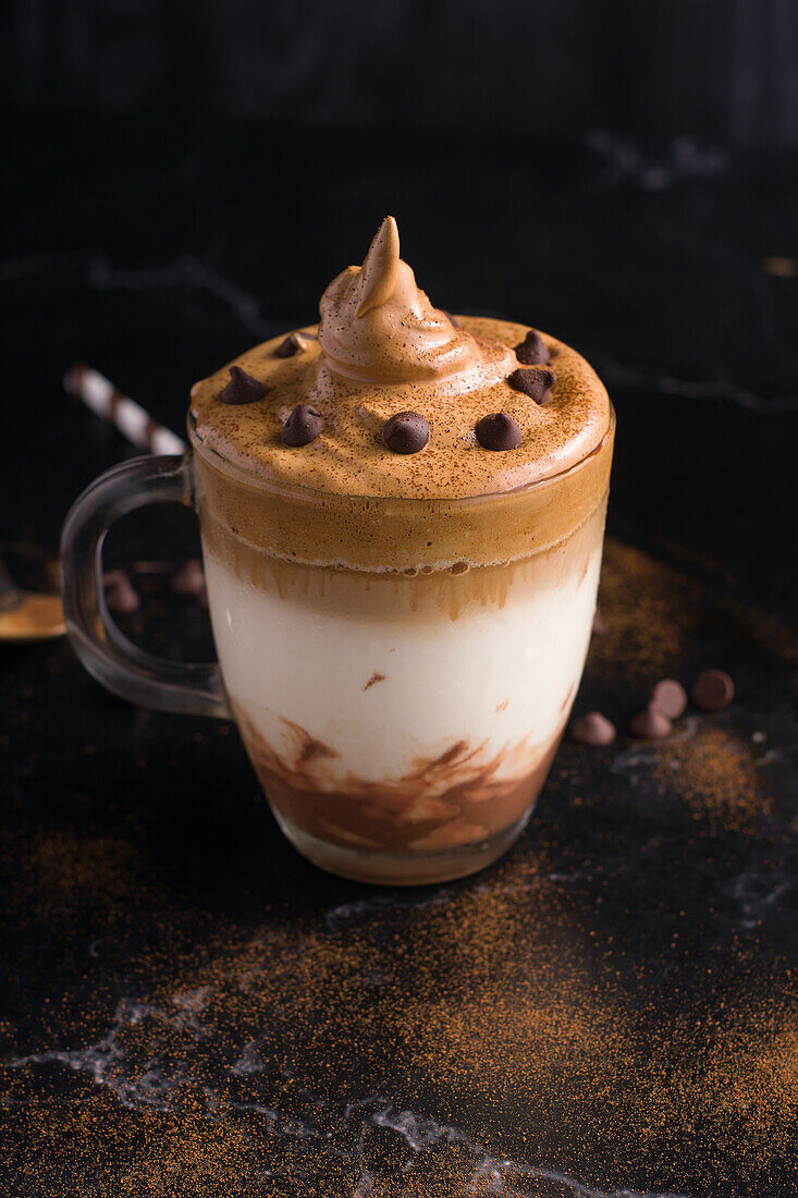 Von oben ein Glas süßer Dalgona-Kaffee mit schaumigem Belag, serviert auf einem Tisch mit Schokoladenwaffelrolle und Kakaopulver