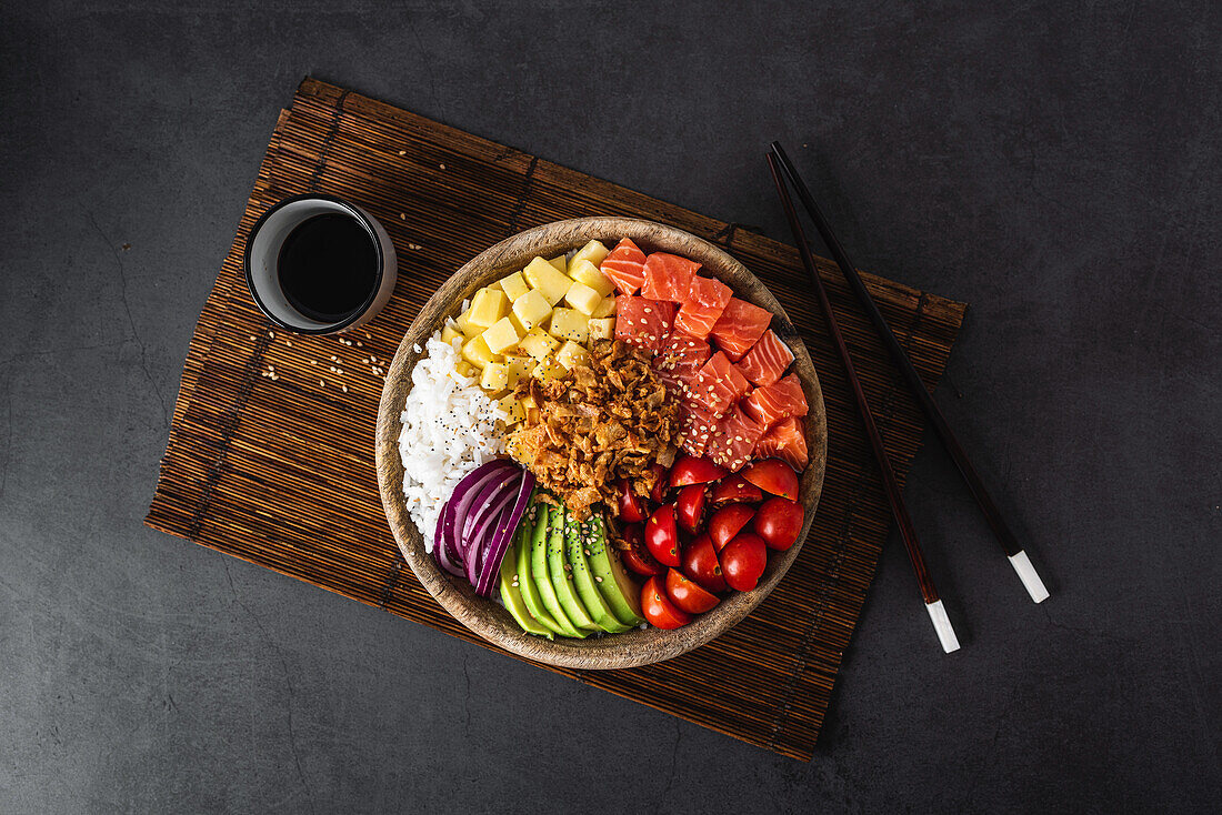 Draufsicht auf eine appetitliche bunte Poke-Schale mit frischem Lachs und Reis, serviert mit reifen Kirschtomaten, Avocadoscheiben, Zwiebeln und Mango auf einem Bambustablett mit Sojasauce