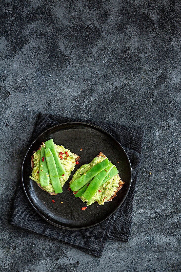 Appetitliche Toasts mit frischer Guacamole und grünen Erbsenschoten garniert auf einem schwarzen Teller