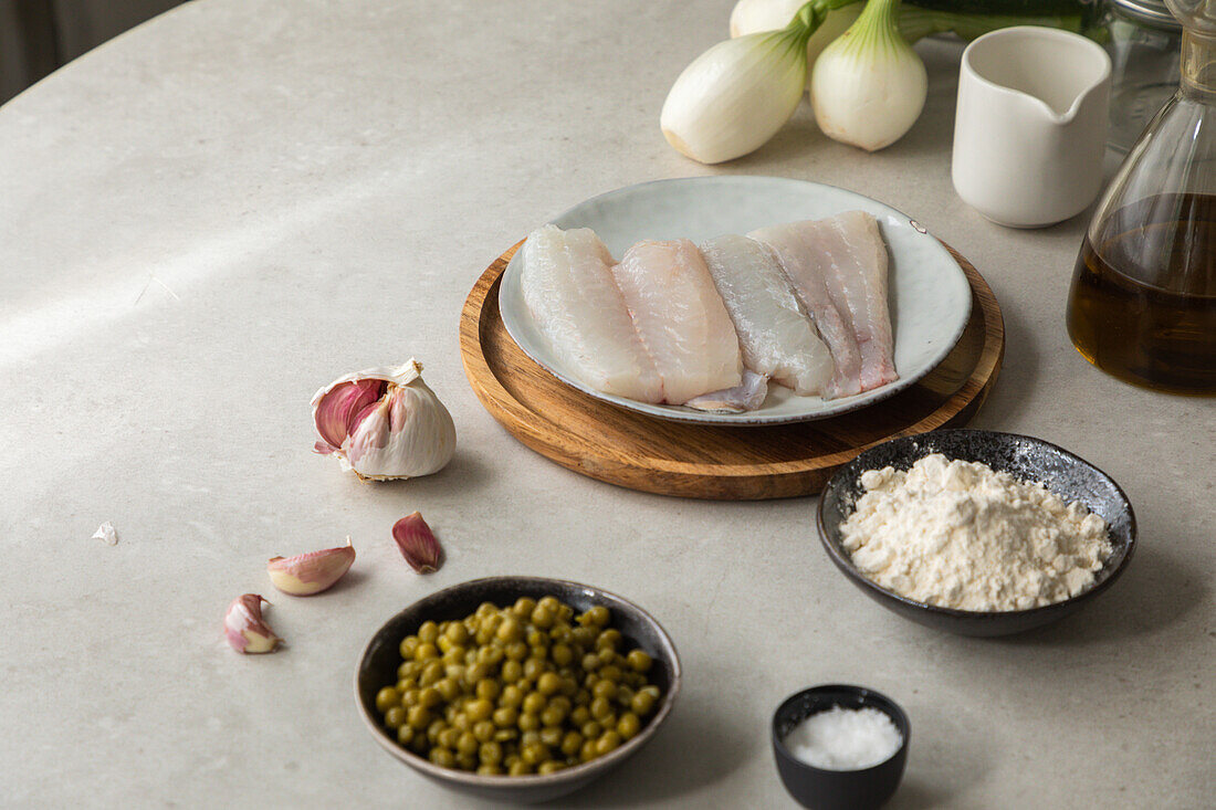 From above fresh garlic cloves and salt placed on table near hake fillet, bowl with peas and flour during food preparation in kitchen