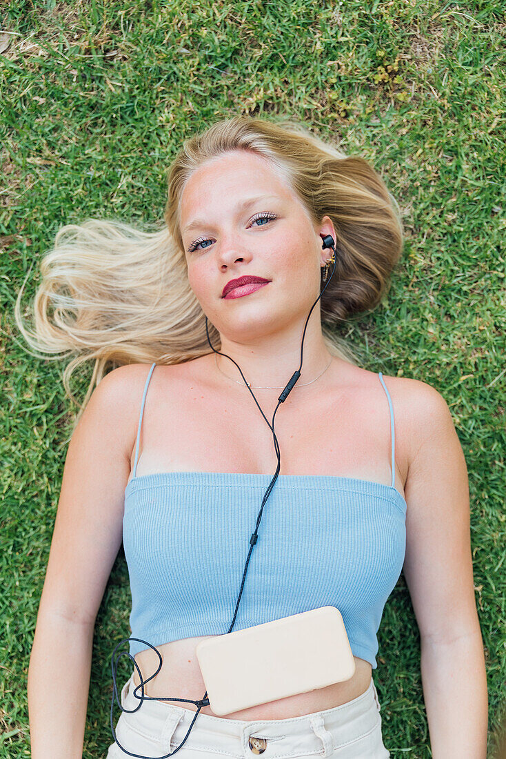 Top view of dreamy female enjoying songs in earphones while lying looking at camera on grass in park in summer