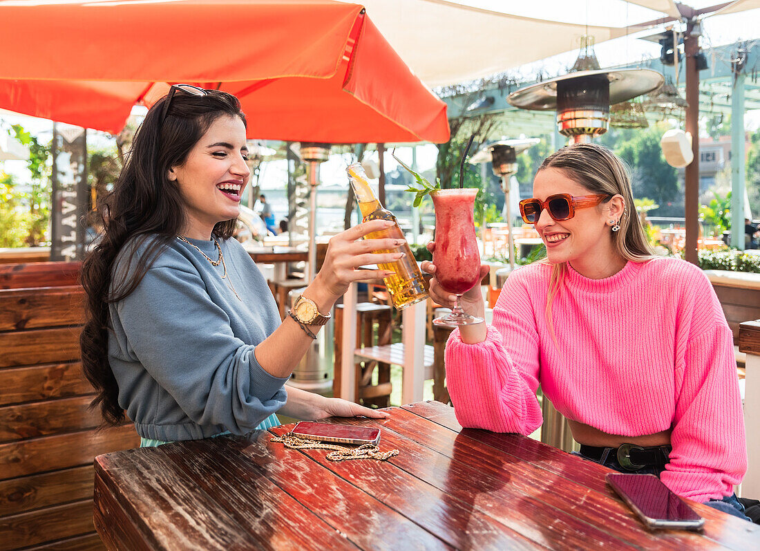 Glückliche junge Freundinnen in legerer Kleidung stoßen mit einer Bierflasche und einem Glas Beerencocktail an, während sie an einem Tisch in einer sonnigen Bar im Freien sitzen