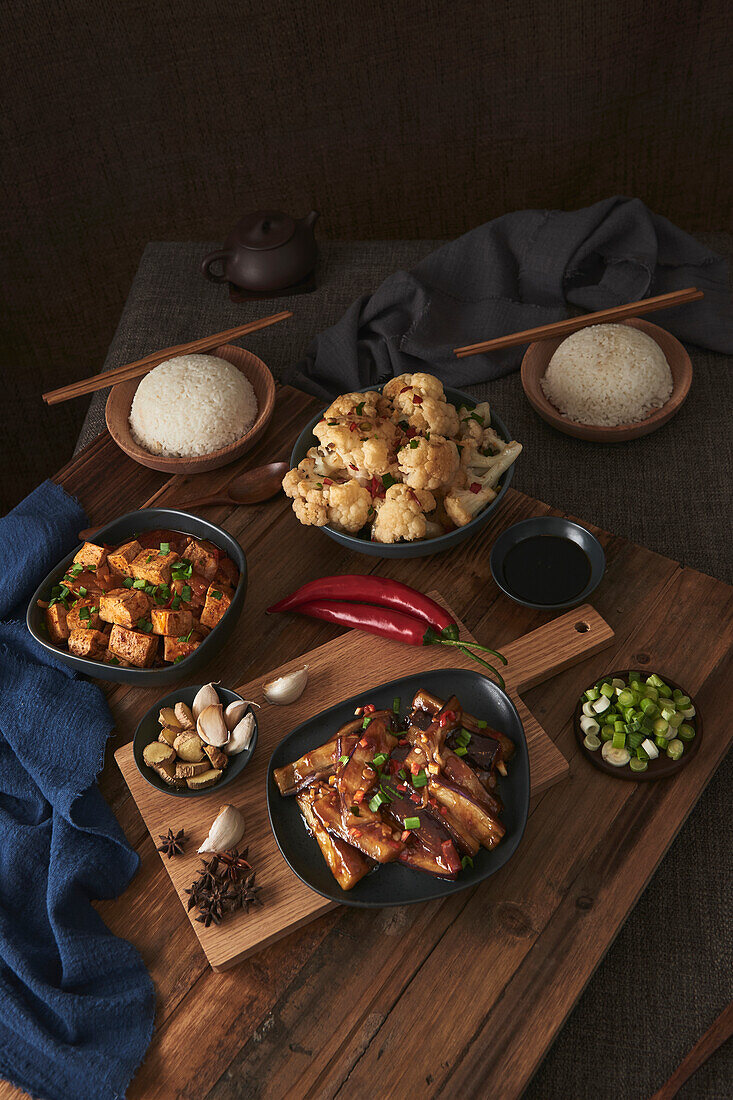 Mala-Tofu und Yuxiang, chinesische vegane Gerichte, begleitet von einer Schüssel mit Reis, Blumenkohl, Sojasauce und einer japanischen Teekanne auf einem mit Stoffen dekorierten Holztisch