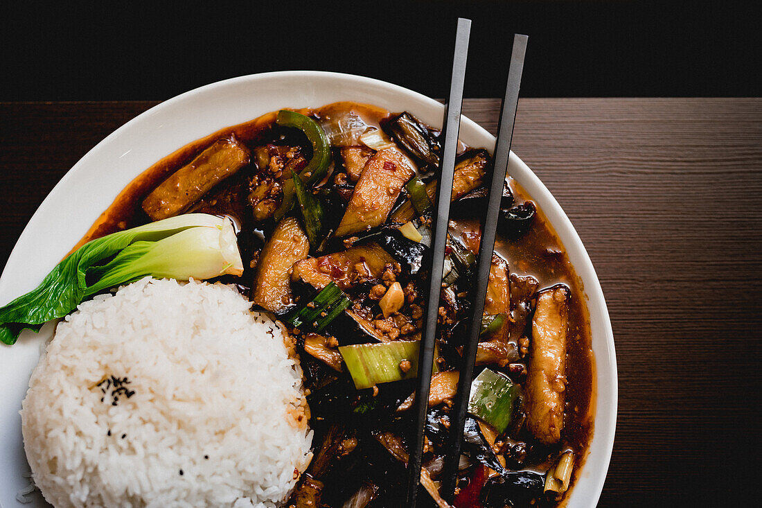 Draufsicht auf appetitlich gekochte Yuxiang-Aubergine mit gesundem Gemüse und Reis auf einem weißen Teller in einem asiatischen Restaurant