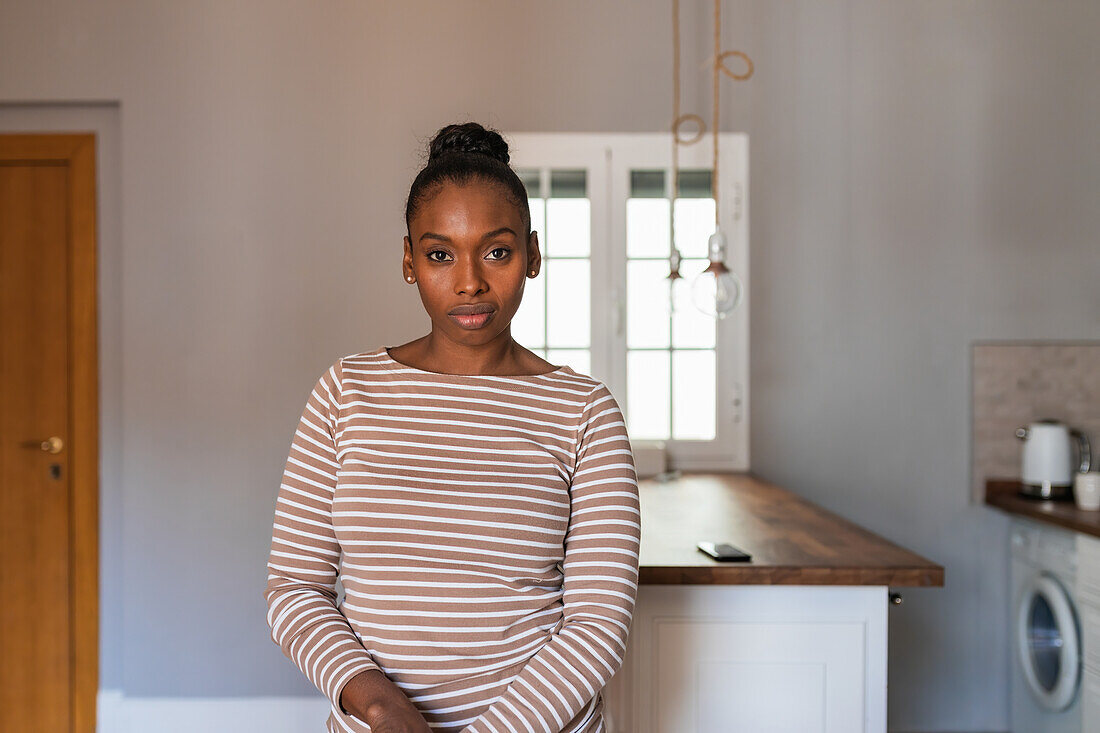 Young African American female with hair bun looking at camera against table and washing machine in kitchen
