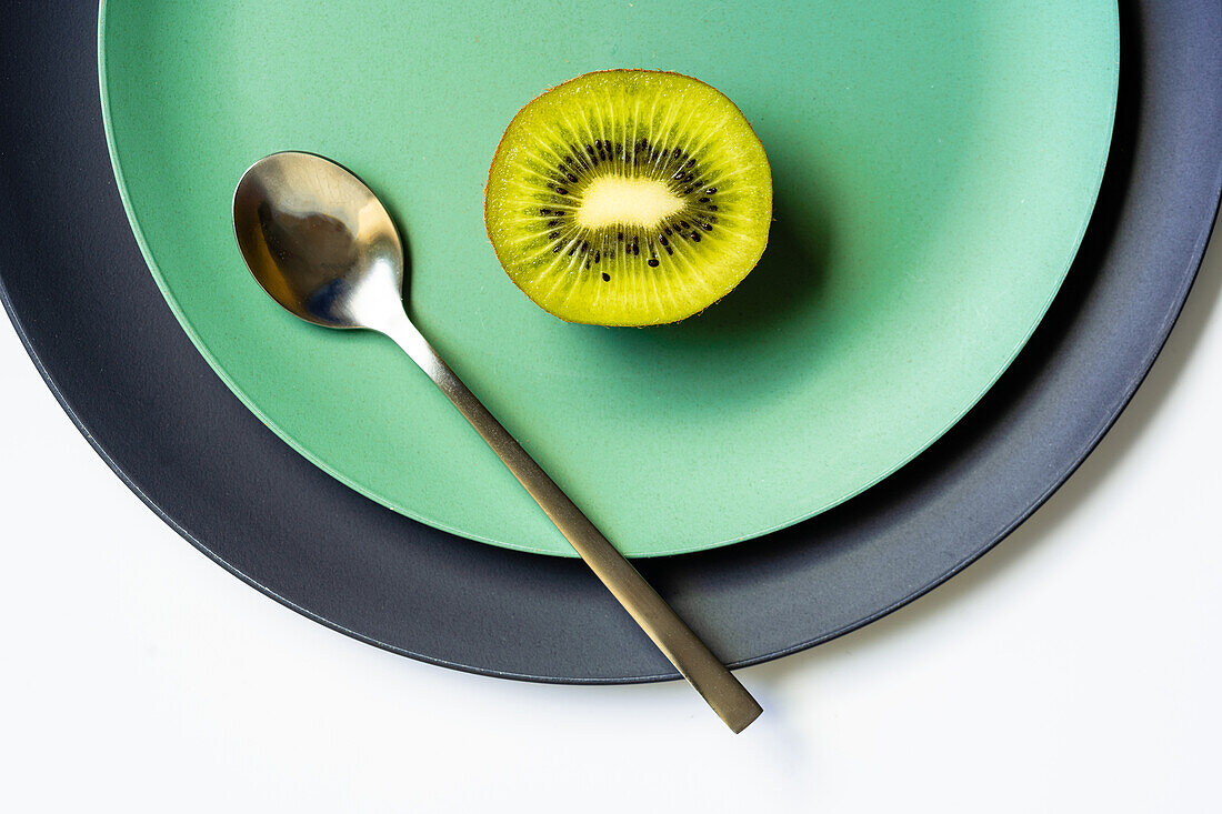 Blick von oben auf die Hälfte einer frischen, reifen, saftigen Kiwi und einen Löffel auf einem grauen und grünen Teller auf einer weißen Fläche