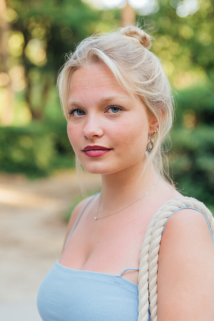 Frau mit blondem Haar steht an einem sonnigen Tag im Sommerpark und schaut in die Kamera