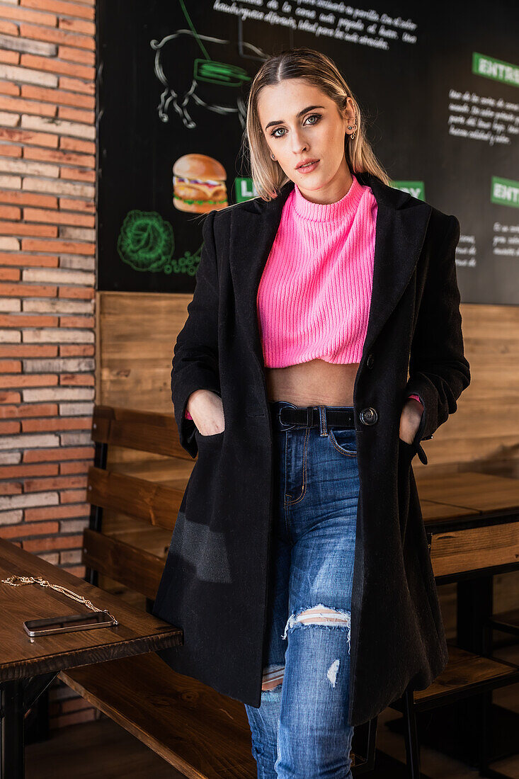 Attraktive junge Frau in Jeans, rosa Pullover und schwarzem Mantel steht mit den Händen in den Taschen in der Nähe eines Tisches im Restaurant und schaut in die Kamera