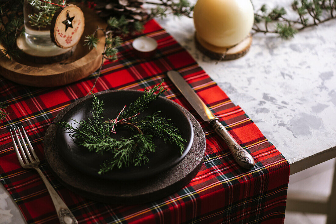 Weihnachtlich gedeckter Tisch mit Kranz auf dem Teller, dekorativem Holzschmuck und rot kariertem Tischtuch mit gelben Lichtern im Hintergrund