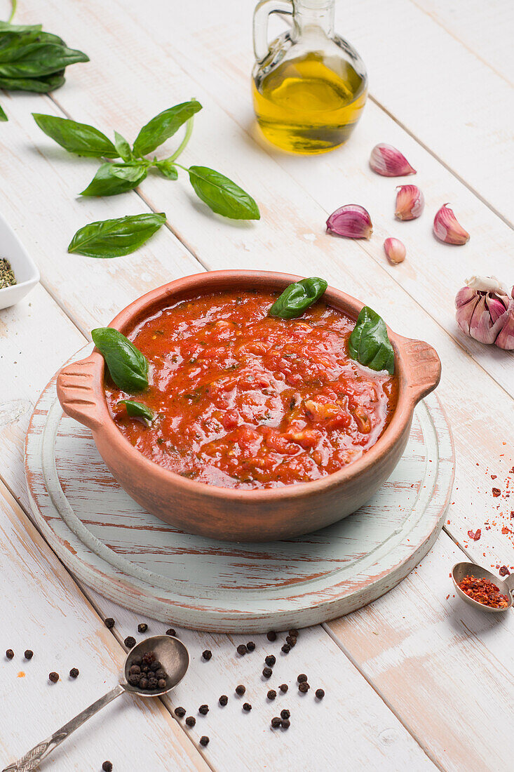Wooden bowl of red marinara sauce made of tomato with basil leaves placed on table with olive oil and garlic