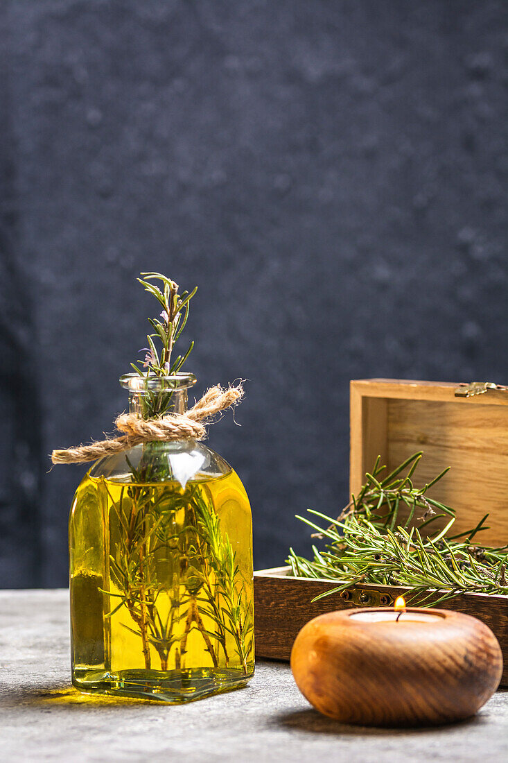 Glasflasche mit ätherischem Öl, Rosmarinzweigen und brennender Bio-Holzkerze auf grauem Tisch