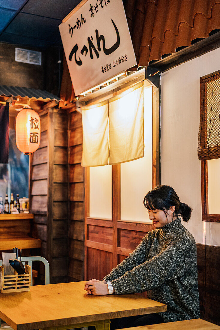 Content Asian woman in casual sweater looking down while sitting at wooden table in ramen bar