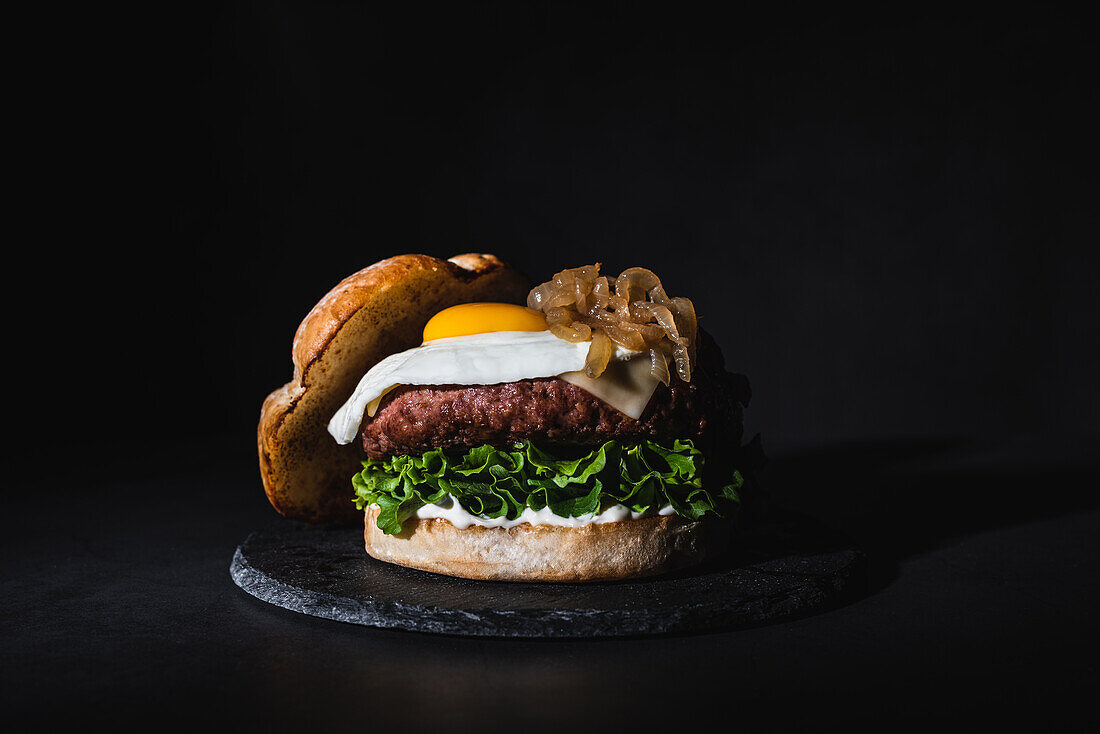 Leckerer Burger mit Ei auf dem Brötchen und frischem Salat auf einer Schiefertafel auf schwarzem Hintergrund im Studio serviert