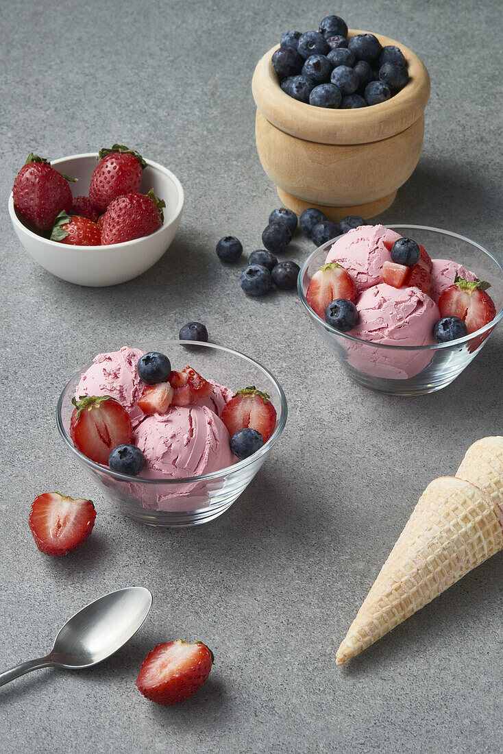 From above of glass bowls of delicious homemade ice cream scoops decorated with fresh strawberries and blueberries and placed on gray table with waffle cones in kitchen