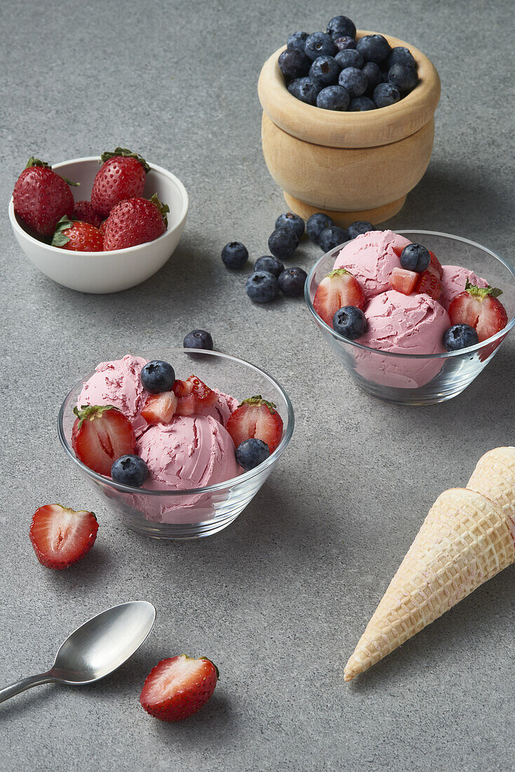 Von oben auf Glasschalen mit köstlichen hausgemachten Eiskugeln, dekoriert mit frischen Erdbeeren und Blaubeeren, auf einem grauen Tisch mit Waffeltüten in der Küche