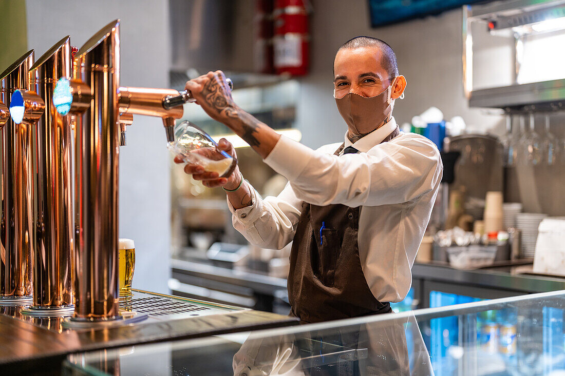 Fröhlicher männlicher Barkeeper in Uniform und Schutzmaske füllt ein Glas mit Bier aus dem Zapfhahn, während er am Tresen einer Kneipe während der Coronavirus-Pandemie arbeitet
