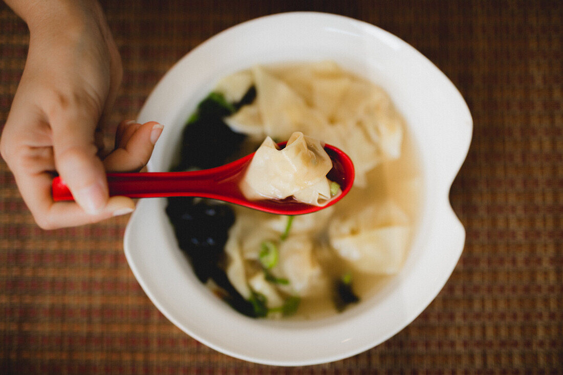 Hand von oben, die eine Schüssel mit leckerer heißer Wonton-Suppe mit rotem Löffel in einem asiatischen Restaurant isst