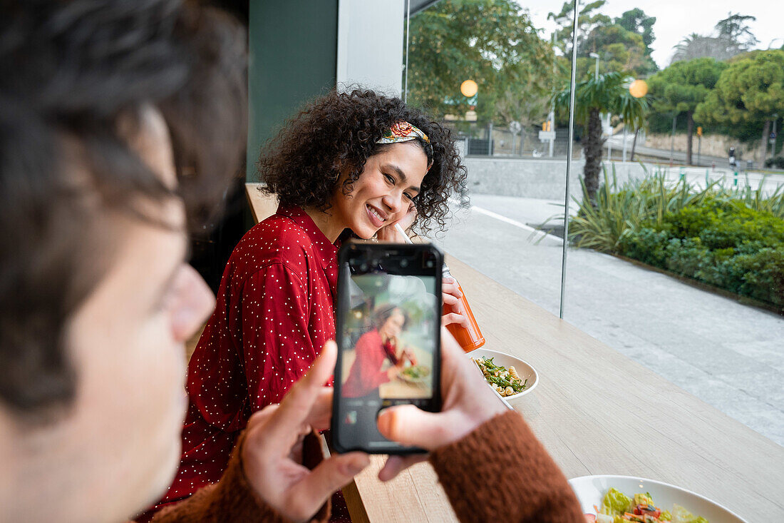 Crop-Mann mit Smartphone fotografiert fröhliche ethnische Freundin beim gesunden Frühstück mit Saft und Salat am Tresen eines modernen Restaurants