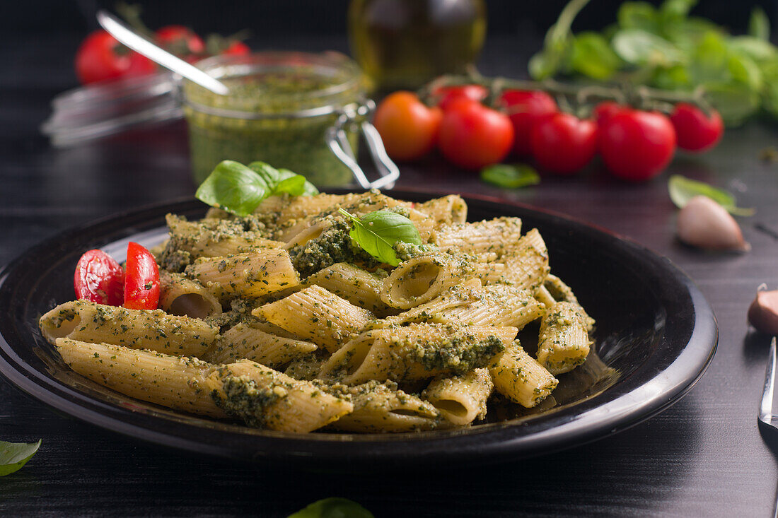 Nahaufnahme eines Tellers mit köstlichen Nudeln mit grüner Pestosauce und Tomaten auf einem schwarzen Holztisch