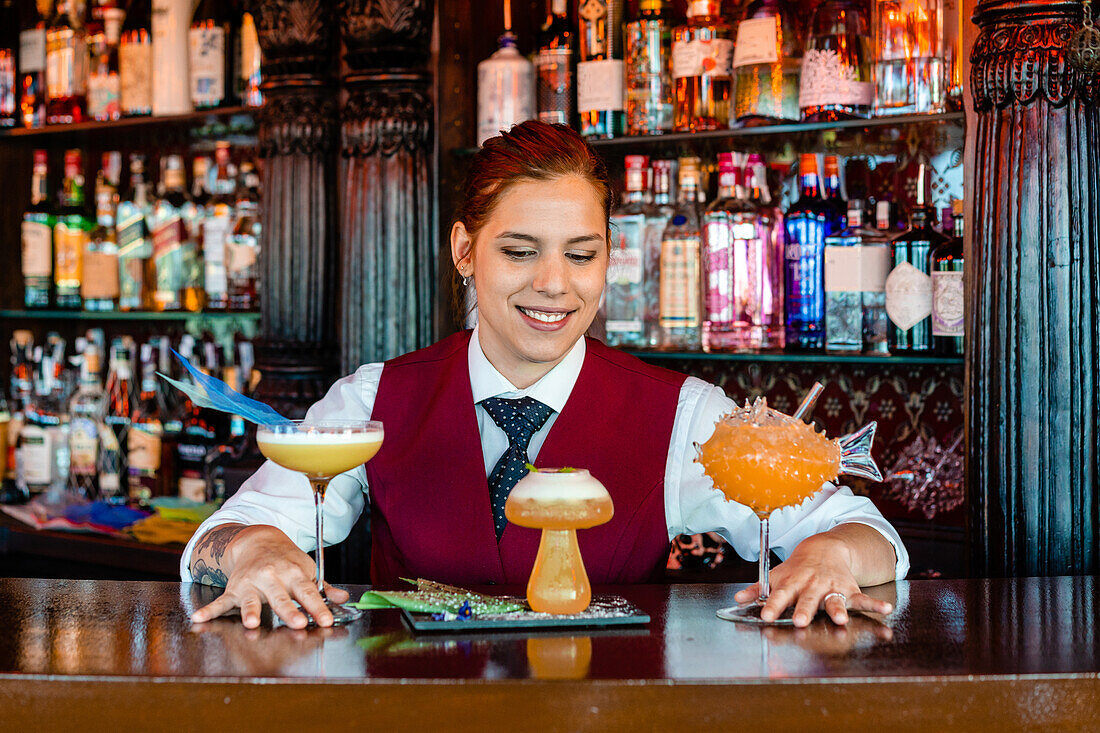 Lächelnde Barkeeperin am Bartresen mit verschiedenen alkoholischen Getränken, die in kreativen Cocktailgläsern in Form von Pilzen und Fischen serviert werden