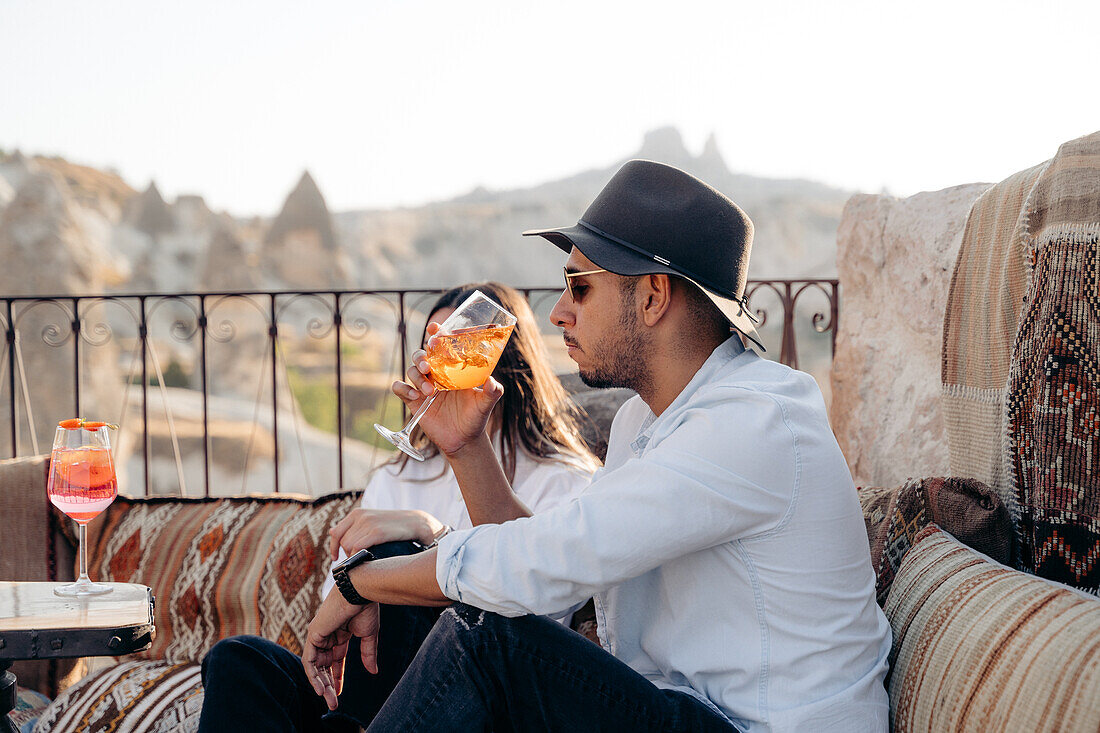 Seitenansicht eines jungen bärtigen Mannes mit Sonnenbrille und modischem Hut, der auf einer Terrasse in Kappadokien, Türkei, einen kalten erfrischenden Cocktail schlürft