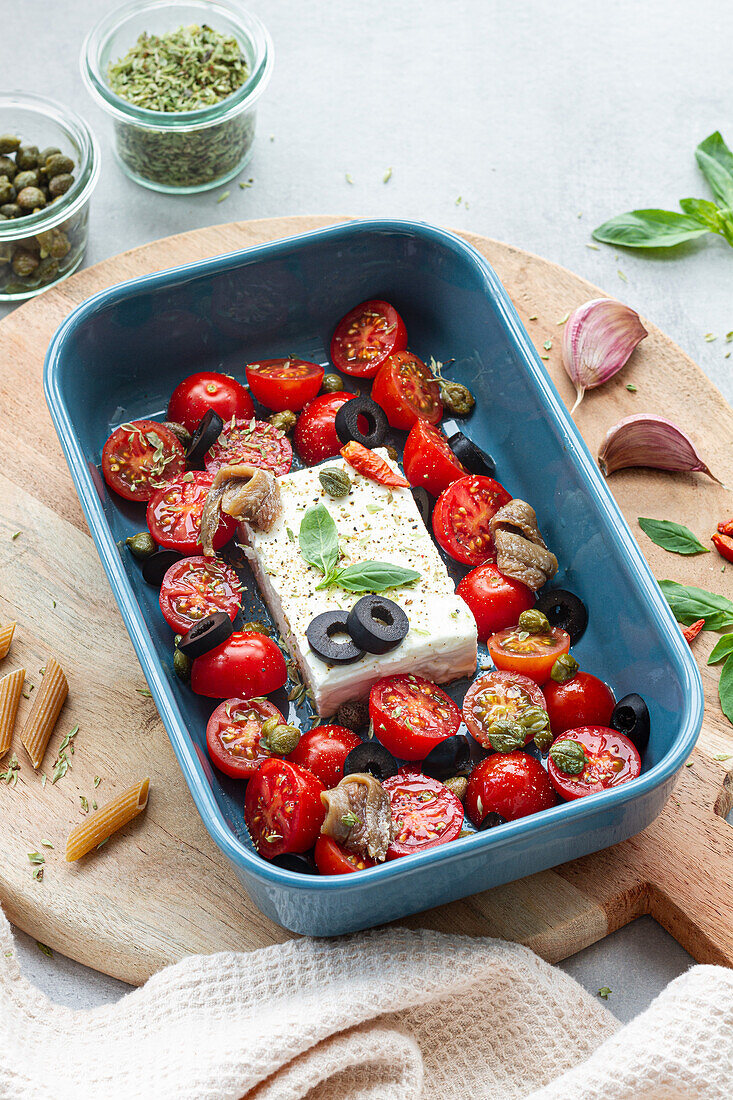 Reife Kirschtomaten mit schwarzen Olivenscheiben und Fetakäse in einer Auflaufform neben ungekochten Penne von oben