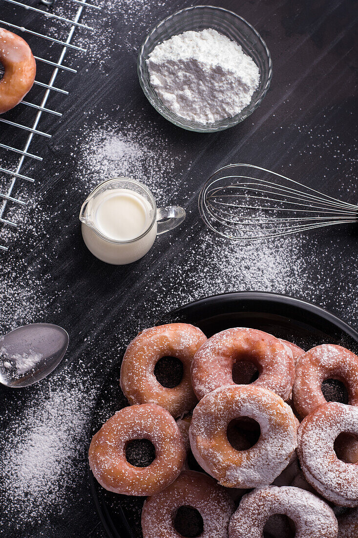 Süße gebratene Krapfen auf einem Teller neben einem Metallkühlregal und einem Milchkännchen auf einem schwarzen, unordentlichen Tisch mit Puderzucker