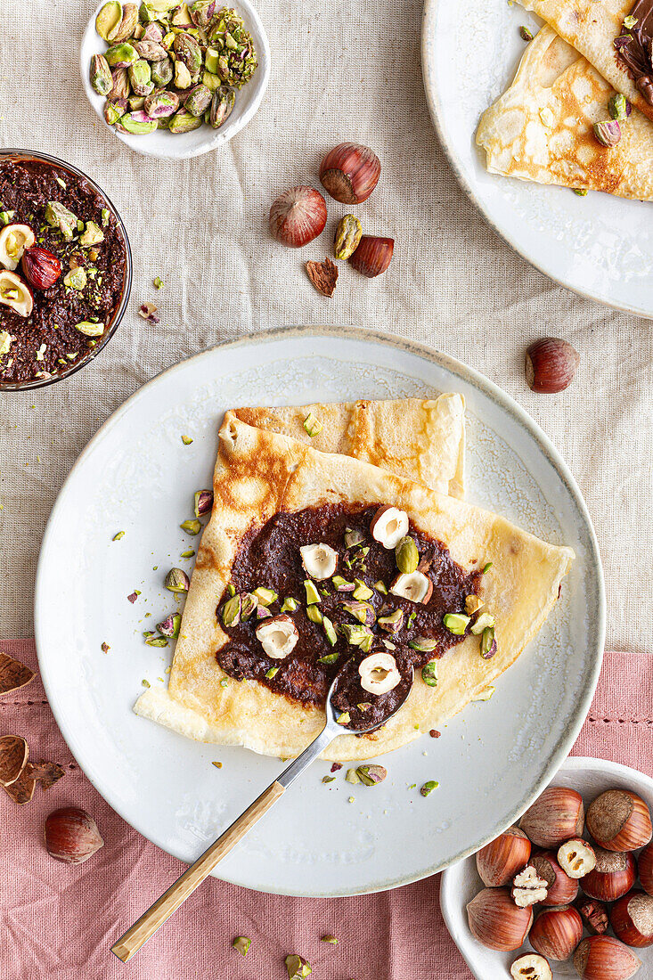 Draufsicht auf leckere Crêpes, die mit Schokolade und Nüssen garniert auf einem Teller auf dem Frühstückstisch serviert werden