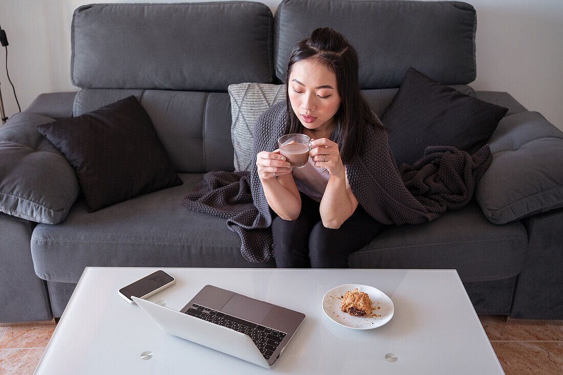 Konzentrierte Asiatin mit Heißgetränk in der Hand schaut auf den Bildschirm eines Netbooks auf dem Tisch mit Gebäck, während sie auf einer bequemen Couch sitzt