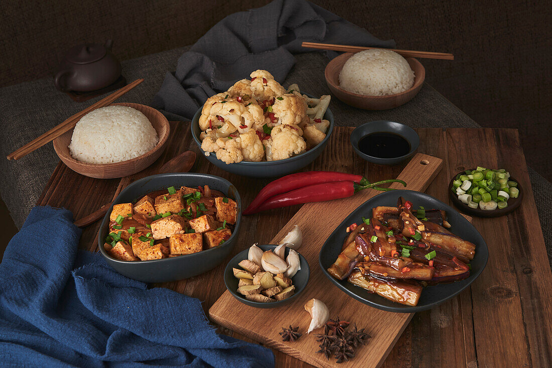 Mala-Tofu und Yuxiang, chinesische vegane Gerichte, begleitet von einer Schüssel mit Reis, Blumenkohl, Sojasauce und einer japanischen Teekanne auf einem mit Stoffen dekorierten Holztisch