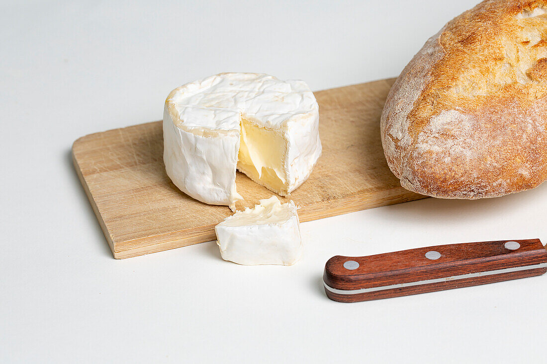 Delicious camembert cheese placed on wooden chopping board near bread and knife on white background
